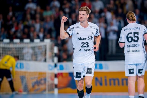 Ein männlicher Handballspieler im weißen Trikot mit der Nummer 23 feiert mit einem Faustschlag auf dem Spielfeld, während ein anderer Mannschaftskamerad mit der Nummer 65 in der Nähe läuft. Der unscharfe Hintergrund zeigt ein überfülltes Stadion und einen Torwart in der Nähe des Tores.