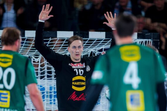 Ein Handballtorwart in Schwarz hebt die Arme vor dem Tor, um einen Schuss abzuwehren. Zwei Spieler in grünen Trikots mit den Nummern 20 und 4 stehen mit dem Rücken zur Kamera im Bildvordergrund.