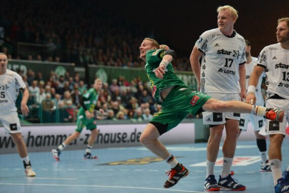 Ein Handballspieler in einem grünen Trikot springt, um den Ball zu schießen, während zwei gegnerische Spieler in weißen Trikots versuchen, ihn während eines überfüllten Hallenspiels zu blockieren.