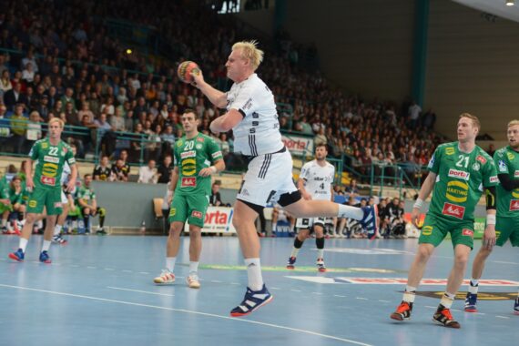 Ein Handballspieler in einem weißen Trikot springt, um den Ball zu werfen, während mehrere gegnerische Spieler in grünen Trikots während eines Spiels in einer mit Zuschauern gefüllten Halle zusehen.