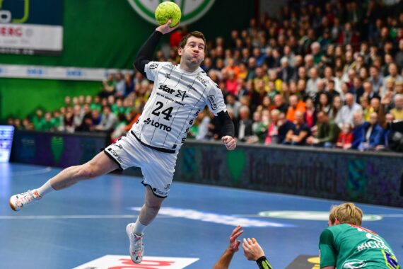 Ein Handballspieler in einem weißen Trikot springt in die Luft, um den Ball zu werfen, während ein Gegner in Grün darunter kauert. Eine Menschenmenge beobachtet das intensive Spiel in einer Halle.
