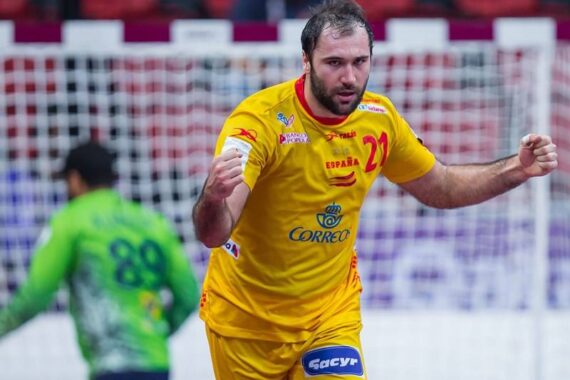 Ein männlicher Handballspieler in einem gelben Trikot jubelt mit geballten Fäusten auf einem Spielfeld, während ein anderer Spieler in einem grünen Trikot in der Nähe des Tores im Hintergrund steht. Die Szenerie scheint ein Hallenspiel zu sein.