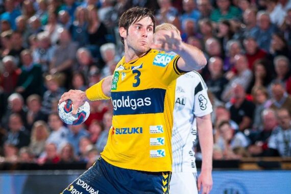 Ein Handballspieler in einem gelb-blauen Trikot bereitet sich während eines Spiels auf den Wurf vor, mit einem gegnerischen Spieler und einer unscharfen Zuschauermenge im Hintergrund.
