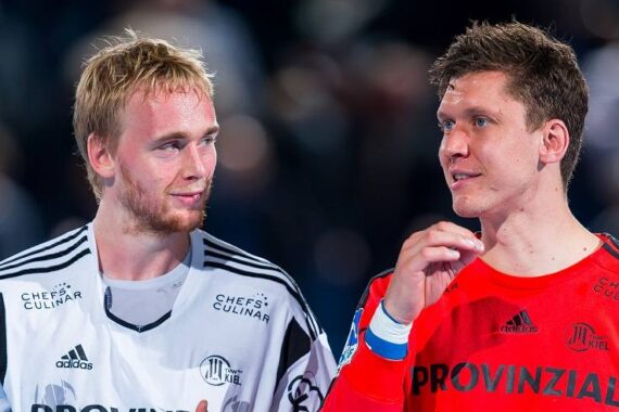 Zwei männliche Sportler in Mannschaftsuniformen, einer in Weiß und einer in Rot, stehen nach einem Spiel eng beieinander, unterhalten sich und lächeln. Der Hintergrund ist unscharf und konzentriert sich auf ihre Interaktion.