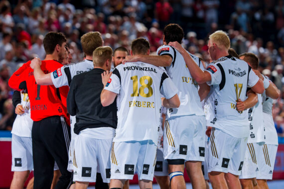 Eine Männer-Handballmannschaft in weißen Trikots kauert auf dem Spielfeld zusammen, die Arme umeinander gelegt, während im Hintergrund eine große Menschenmenge zusieht. Der Spieler in der Mitte trägt das Trikot mit der Nummer 18.