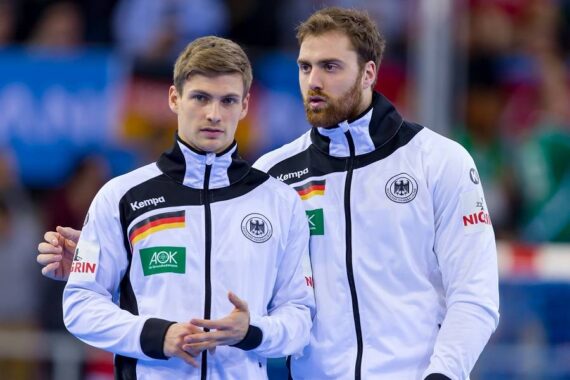 Zwei männliche Sportler in weißen Jacken der deutschen Nationalmannschaft stehen nebeneinander. Der eine blickt nach vorne, während der andere mit ihm spricht. Der Hintergrund ist unscharf und zeigt eine Sporthalle und Zuschauer.