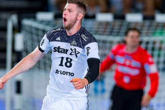 Ein männlicher Handballspieler in einem weiß-schwarzen Trikot mit der Nummer 18 jubelt ausgelassen auf dem Spielfeld, während im Hintergrund in der Nähe des Tores ein verschwommener Torwart in Rot zu sehen ist.