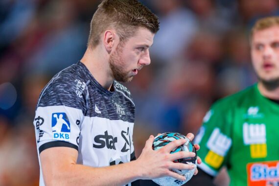 Ein Handballspieler in einem weiß-schwarzen Trikot hält einen blau-schwarzen Handball und bereitet sich auf den Wurf vor, während im Hintergrund ein anderer Spieler in einem grünen Trikot zu sehen ist. Die Szene spielt in einer Sporthalle.