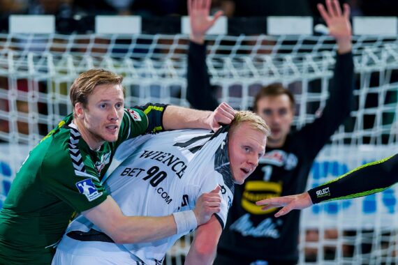 Zwei männliche Handballspieler kämpfen in der Nähe des Tores um eine Position, wobei der eine in einem grünen Trikot das Trikot seines Gegners in einem weißen Trikot ergreift. Im Hintergrund sind ein Torwart und ein Tornetz zu sehen.