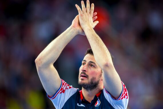 Ein männlicher Handballspieler in einem blau-roten Trikot steht mit über dem Kopf verschränkten Händen da und blickt nach oben, während im Hintergrund eine verschwommene Menschenmenge zu sehen ist.