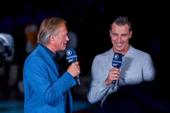 Zwei Männer stehen und sprechen während eines Interviews in Mikrofone. Einer trägt einen blauen Blazer, der andere einen hellgrauen Pullover und lächelt. Beide halten Mikrofone mit blauen Abdeckungen mit weißen Logos. Der Hintergrund ist dunkel.