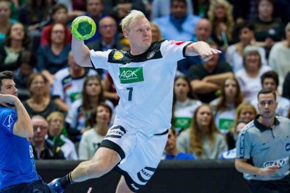 Ein männlicher Handballspieler in einem weiß-schwarzen Trikot springt in der Luft, um den Ball zu werfen, während ein Gegner in Blau und die Schiedsrichter zusehen. Eine Zuschauermenge füllt den Hintergrund.