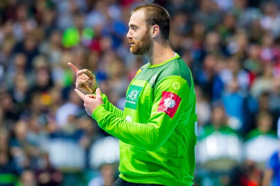 Ein Handballtorwart in einem hellgrünen Trikot gestikuliert während eines Spiels mit den Händen, während im Hintergrund die Zuschauer und Spieler verschwommen zu sehen sind.