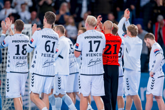 Eine Gruppe männlicher Handballspieler in weißen Trikots mit schwarzen Nummern und einer in einem roten Trikot stehen mit dem Gesicht zum Spielfeld und heben die Hände, während im Hintergrund ein Publikum zusieht.