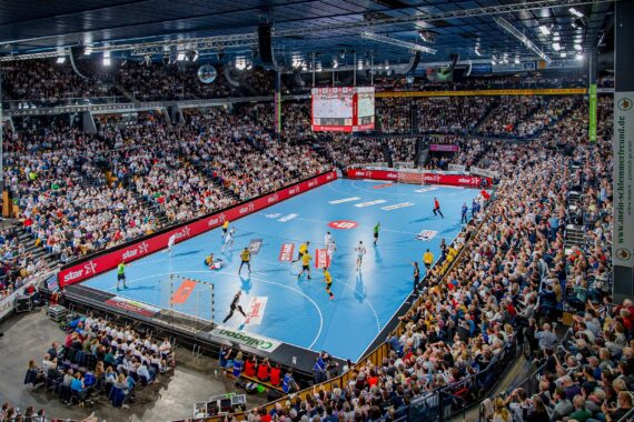 Eine überfüllte Halle mit Tausenden von Zuschauern verfolgt ein professionelles Handballspiel auf einem leuchtend blauen Spielfeld, mit Spielern in Aktion und digitalen Anzeigetafeln über dem Spielfeld.