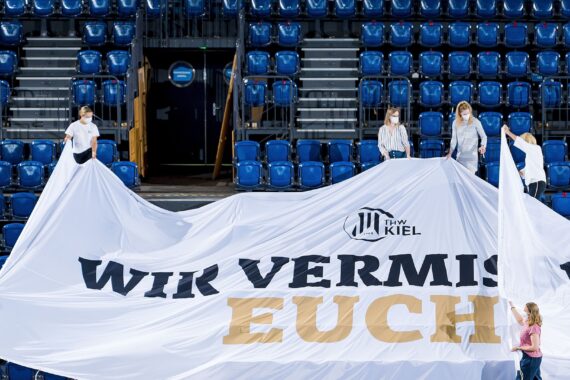 Vier Personen halten ein großes weißes Transparent in einem leeren Stadion mit blauen Sitzen. Auf dem Transparent steht THW Kiel WIR VERMISSEN EUCH.