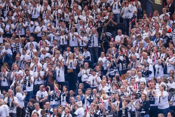Eine große Menge enthusiastischer Sportfans, viele mit weißen Hemden und Mannschaftsschals, jubeln und klatschen auf den Sitzen im Stadion und schaffen eine lebhafte und energiegeladene Atmosphäre.