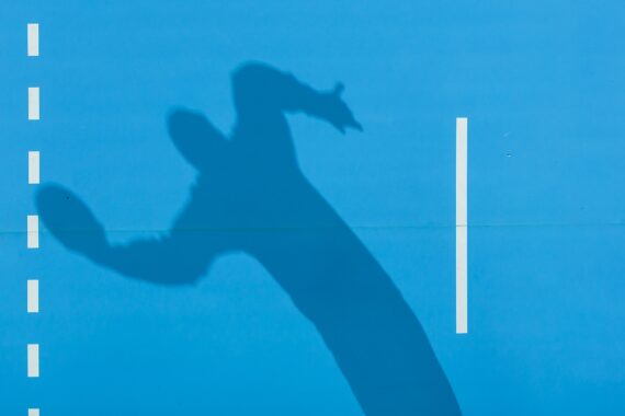 Der Schatten einer Person, die Tennis spielt und einen Schläger in Bewegung hält, wird auf ein hellblaues Spielfeld geworfen, auf dem weiße gestrichelte und durchgezogene Linien sichtbar sind.