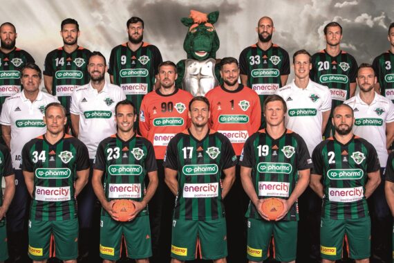 Eine Herren-Handballmannschaft in grün-schwarzen Trikots posiert für ein Gruppenfoto und hält orangefarbene Bälle in der Hand. Trainer und Betreuer in weißen Hemden stehen dahinter, zusammen mit einem grünen Drachenmaskottchen. Ein bewölkter Himmel bildet den Hintergrund.