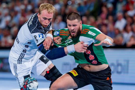 Zwei männliche Handballspieler in Aktion; der eine in einem weiß-schwarzen Trikot verteidigt gegen den anderen in einem grün-roten Trikot, beide zeigen intensive Mienen während eines Spiels, mit einer unscharfen Menge im Hintergrund.