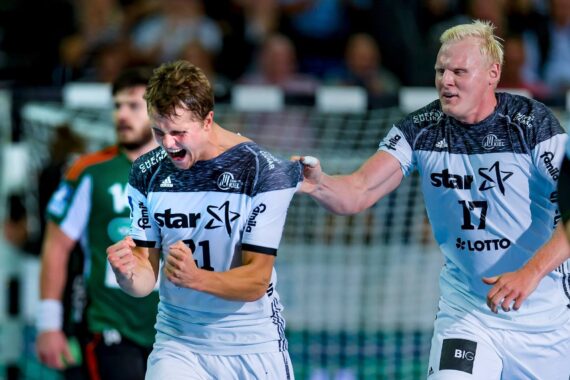 Zwei männliche Handballspieler in passenden schwarz-weißen Trikots feiern ausgelassen auf dem Spielfeld; einer ballt die Fäuste und schreit, während der andere lächelt und sich an die Schulter fasst. Im Hintergrund ist ein Torpfosten zu sehen.