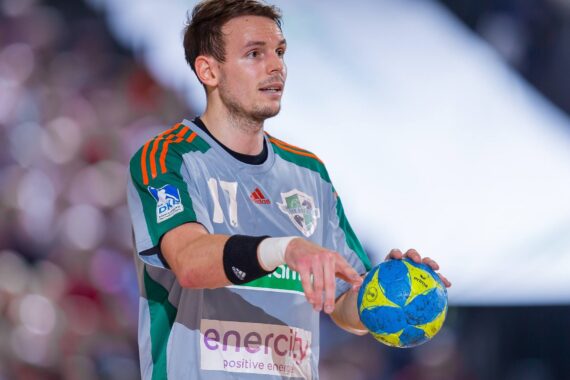Ein männlicher Handballspieler in einem grau-grünen Trikot hält einen blau-gelben Handball und wirkt während eines Spiels konzentriert. Der Hintergrund ist unscharf und hebt den Spieler hervor.