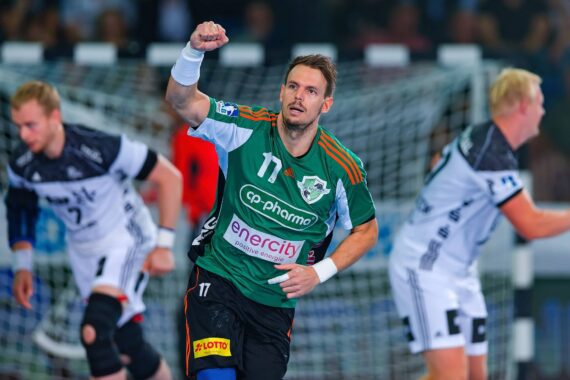 Ein Handballspieler in einem grün-schwarzen Trikot hebt während eines Spiels jubelnd die Faust, während im Hintergrund zwei gegnerische Spieler in weißen und schwarzen Trikots zu sehen sind.