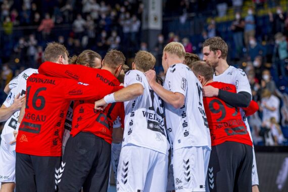 Eine Gruppe männlicher Handballspieler in rot-weißen Trikots drängt sich auf einem Hallenplatz zusammen, während im Hintergrund eine unscharfe Menschenmenge zu sehen ist.