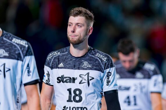 Ein männlicher Sportler in einem weiß-schwarzen Mannschaftstrikot mit der Nummer 18 steht auf einem Spielfeld und blickt nach vorne. Andere Spieler in ähnlichen Trikots sind im Hintergrund zu sehen. Der Schauplatz scheint eine Hallensportveranstaltung zu sein.