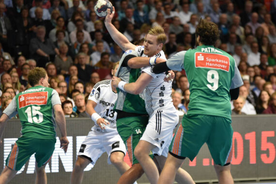 Zwei Mannschaften liefern sich ein intensives Handballspiel; ein Spieler in Weiß versucht, sich zu befreien, während er von drei Spielern in grünen Trikots eng verteidigt wird, während die Zuschauer auf der Tribüne zusehen.