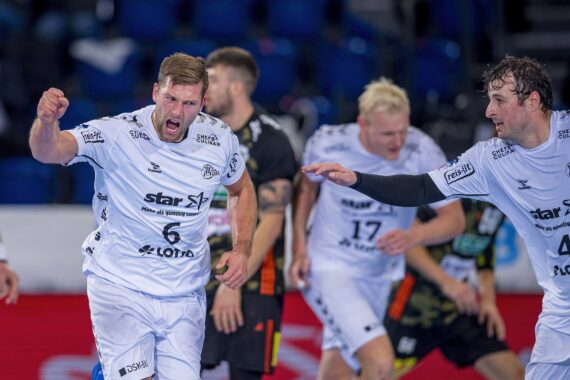Männliche Handballspieler in weißen Trikots feiern auf einem Hallenplatz mit entschlossenem Gesichtsausdruck und geballten Fäusten, während im Hintergrund gegnerische Spieler in dunklen Trikots stehen.
