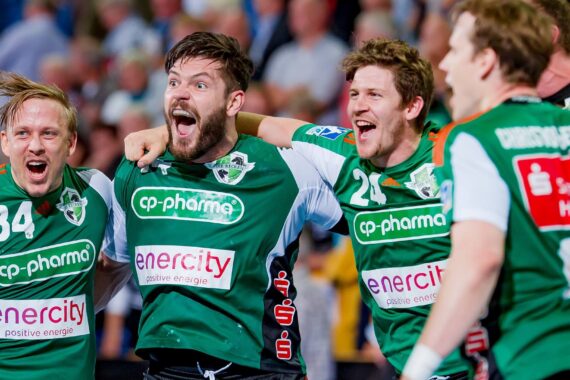 Vier männliche Handballspieler in grünen Trikots feiern ausgelassen, lächelnd und schreiend, die Arme umeinander gelegt, vor einer jubelnden Menge.