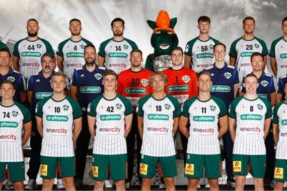Eine Handballmannschaft posiert für ein Gruppenfoto in Uniformen, mit Trainern auf beiden Seiten und einem Maskottchen in der hinteren Reihe. Die Kulisse scheint ein Stadion mit einem dramatischen, bewölkten Himmel im Hintergrund zu sein.