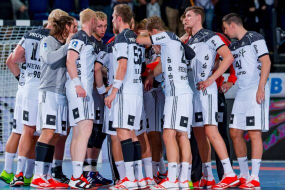 Eine Herren-Handballmannschaft in weiß-schwarzen Trikots versammelt sich auf dem Spielfeld und hört ihrem Trainer vor einem Spiel oder während einer Auszeit zu. Die Spieler sind konzentriert und engagiert bei der Diskussion.