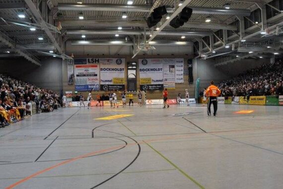 Eine überfüllte Sporthalle mit Zuschauern auf der Tribüne, die ein Handballspiel verfolgen; die Spieler sind auf dem Spielfeld zu sehen, und an der gegenüberliegenden Wand ist Werbung angebracht.