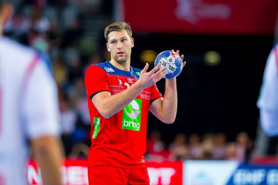 Ein männlicher Handballspieler in einem roten Trikot hält einen blau-weißen Ball und bereitet sich auf einen Pass oder einen Schuss während eines Spiels vor. Andere Spieler in weißen Trikots sind im Vorder- und Hintergrund zu sehen.