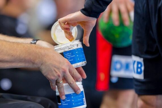 Eine Nahaufnahme von zwei Personen, die Handcreme aus einem Plastiktiegel verwenden. Eine Person hält den Tiegel, während die andere die Finger in die Creme taucht. Im Hintergrund sind ein verschwommener grüner Handball und Sportuniformen zu sehen.