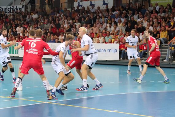 Eine Gruppe von Männern spielt in einer Halle ein intensives Handballspiel, bei dem Spieler in rot-weißen Trikots um den Ball kämpfen. Auf den Tribünen im Hintergrund sitzen Zuschauer, die das Geschehen aufmerksam verfolgen.