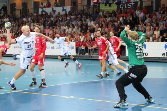 Ein Handballspieler in einem weißen Trikot bereitet sich darauf vor, den Ball auf das Tor zu werfen, während ein Torwart in Grün den Ball abwehrt. Mehrere Spieler in Rot und eine große Menschenmenge verfolgen das Geschehen in einer Halle.