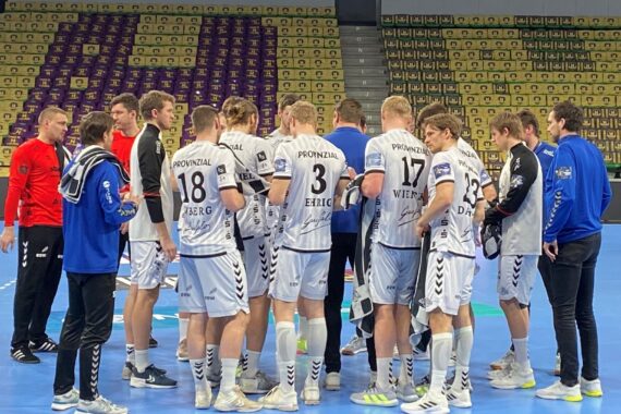 Eine Herren-Handballmannschaft in weißen Trikots kauert auf einem blauen Hallenplatz zusammen, mit Trainern in Blau und Rot. Gelbe und lila Sitze füllen die leeren Tribünen im Hintergrund.