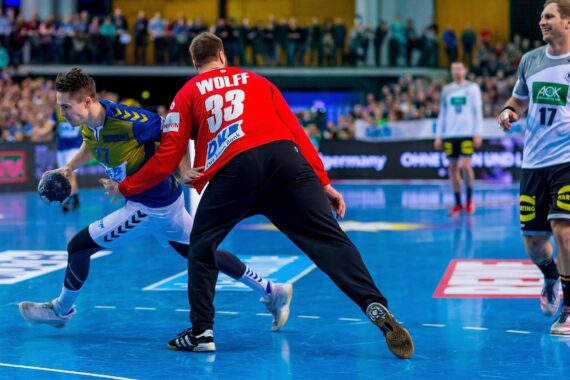 Ein Handballspieler in einem blau-gelben Trikot versucht, einem Torwart in einem roten Trikot auszuweichen, während andere Spieler und Zuschauer im Hintergrund zusehen. Die Szene spielt sich auf einem hellblauen Hallenplatz ab.