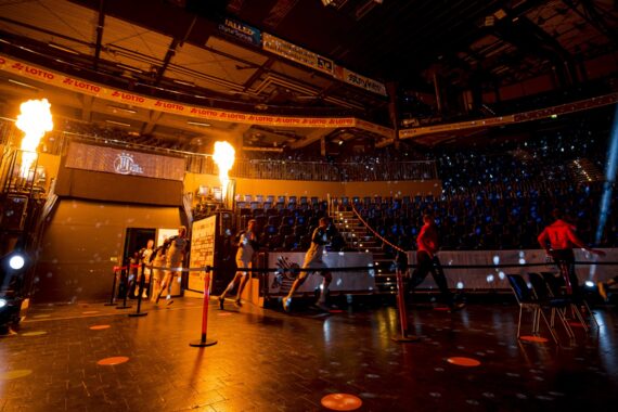 Die Athleten betreten eine hell erleuchtete Sportarena, in der auf beiden Seiten des Eingangs Flammen emporschießen und im Hintergrund leere blaue Sitzreihen zu sehen sind. Die Atmosphäre ist dramatisch und energiegeladen.