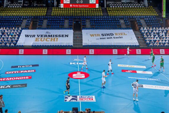 In einer hell erleuchteten Halle ohne Fans wird ein Handballspiel ausgetragen; auf den Tribünen hängen Transparente mit den Aufschriften Wir vermissen euch! und Wir sind Kiel ... nur mit euch! auf Deutsch. Die Spieler stehen auf einem blauen Spielfeld.