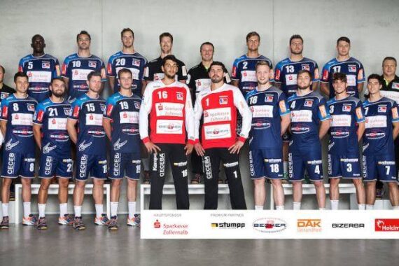 Eine Herren-Handballmannschaft posiert in einer Halle vor einer grauen Wand. Die Spieler tragen blaue Trikots, mit Ausnahme von zwei Torhütern in roten Trikots, und werden von Trainern und Mitarbeitern in weißen Hemden begleitet. Vor der Gruppe ist ein Sponsorenbanner angebracht.