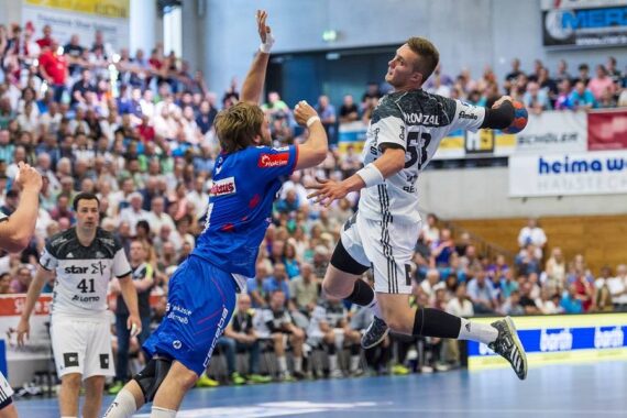 Zwei männliche Handballspieler befinden sich während eines intensiven Spiels in der Luft, wobei der eine in Blau einen Schussversuch eines Spielers in Weiß abwehrt. Die Zuschauer schauen von der überfüllten Tribüne im Hintergrund zu.