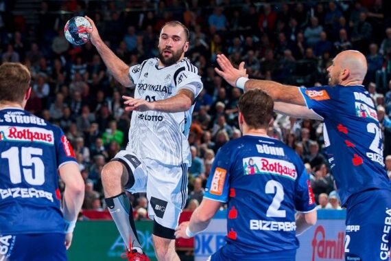 Ein Handballspieler in einem weißen Trikot bereitet sich auf einen Sprungwurf vor, während zwei Verteidiger in Blau versuchen, ihn zu stoppen. Die Zuschauer im Hintergrund verfolgen das intensive Geschehen.