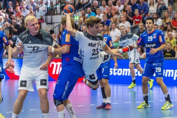 Ein Handballspieler in einem weißen Trikot springt, um den Ball zu schießen, während er von zwei Spielern in blauen Trikots während eines Spiels verteidigt wird, während die Zuschauer im Hintergrund zuschauen.