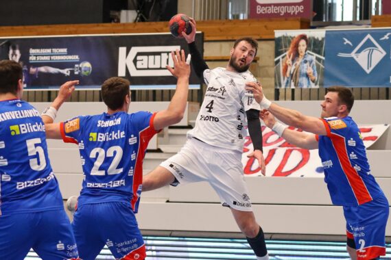 Ein Handballspieler in einem weißen Trikot springt zum Schuss, während drei Verteidiger in blauen Trikots versuchen, ihn während eines Hallenspiels zu blockieren. Im Hintergrund sind das Spielfeld und die Fahnen zu sehen.