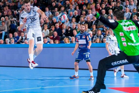 Ein Handballspieler in einem weißen Trikot springt und schießt in Richtung Tor, während der Torwart in Grün sich darauf vorbereitet, das Tor zu blockieren. Zwei Spieler in Blau schauen zu, während eine Menschenmenge den Hintergrund füllt.