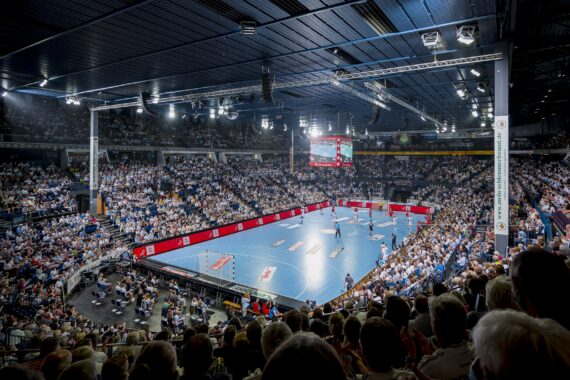 Eine große, hell erleuchtete Halle mit vielen Zuschauern, die ein Handballspiel auf einem blauen Spielfeld verfolgen, mit digitalen Anzeigetafeln und Werbung rund um den Spielbereich.